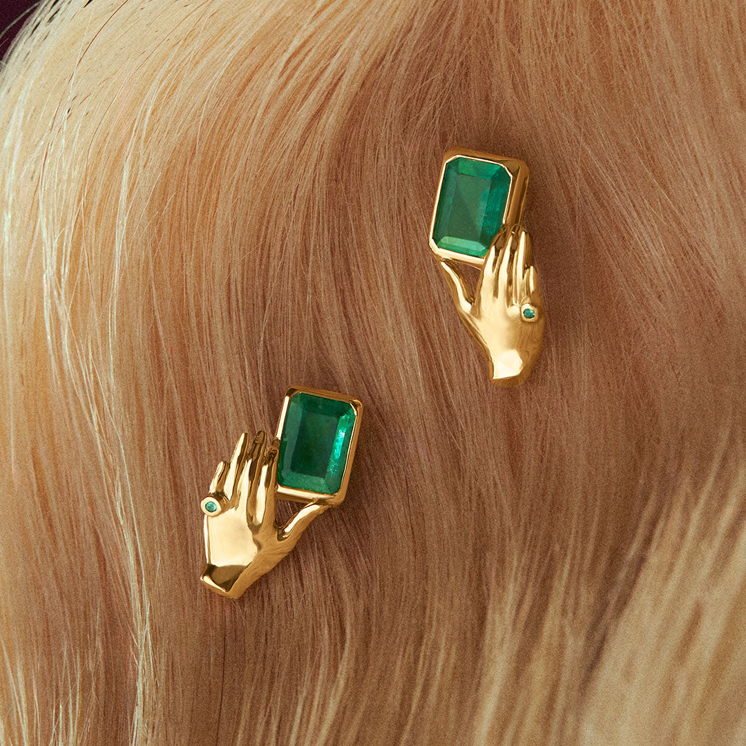 Brincos de ouro 18k de mãos segurando esmeralda retangular